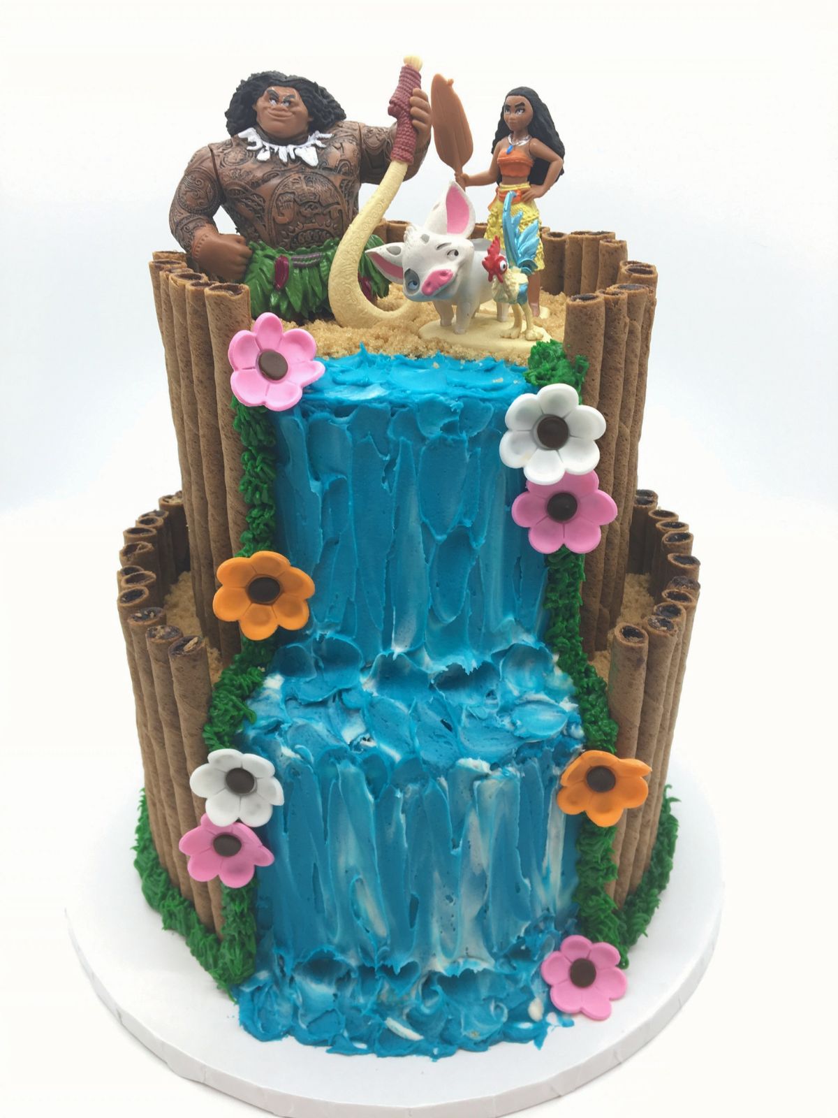 A Moana Birthday Cake with two tiers and a buttercream waterfall with Pirouline stick cookies and fondant flowers, and a Maui and Moana cake topper.